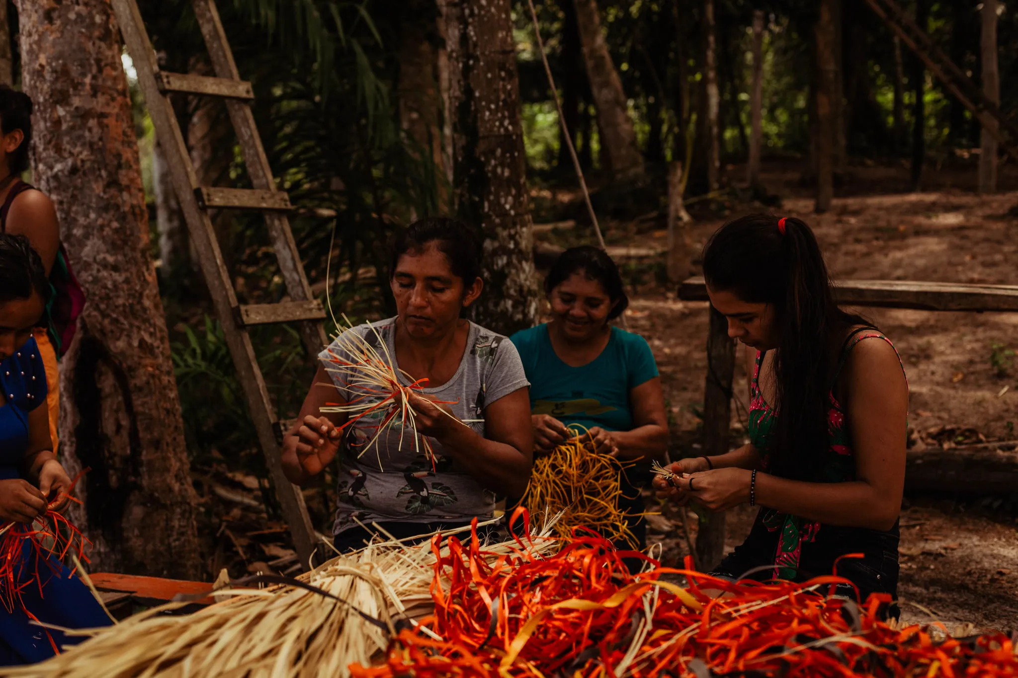 Cinco mujeres de la comunidad sentadas en el suelo entre los árboles, trenzando haces de paja teñidos de naranja y rojo