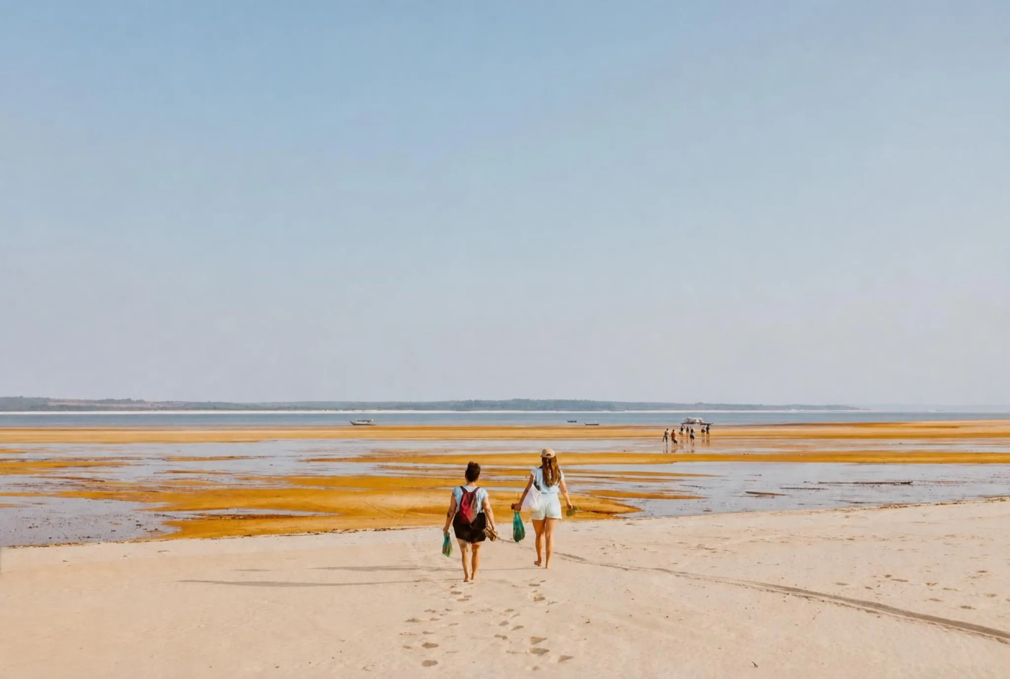 Dos personas alejándose a través de un enorme banco de arena pálida, con más figuras y botes amarrados a lo lejos, en la línea del agua, bajo un cielo brumoso