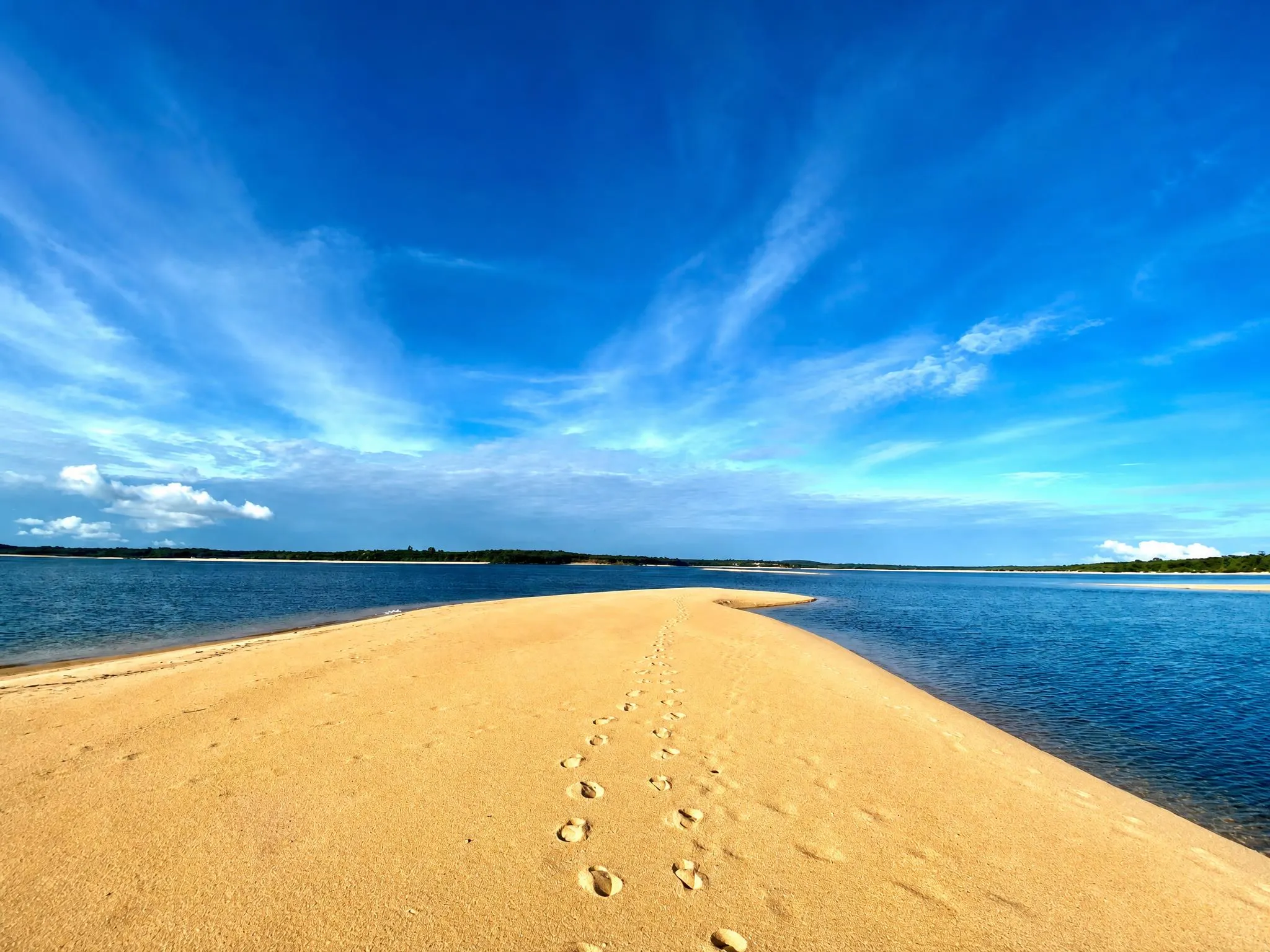 A golden sandbank with a trail of footprints, cutting through blue water on both sides, under an open sky