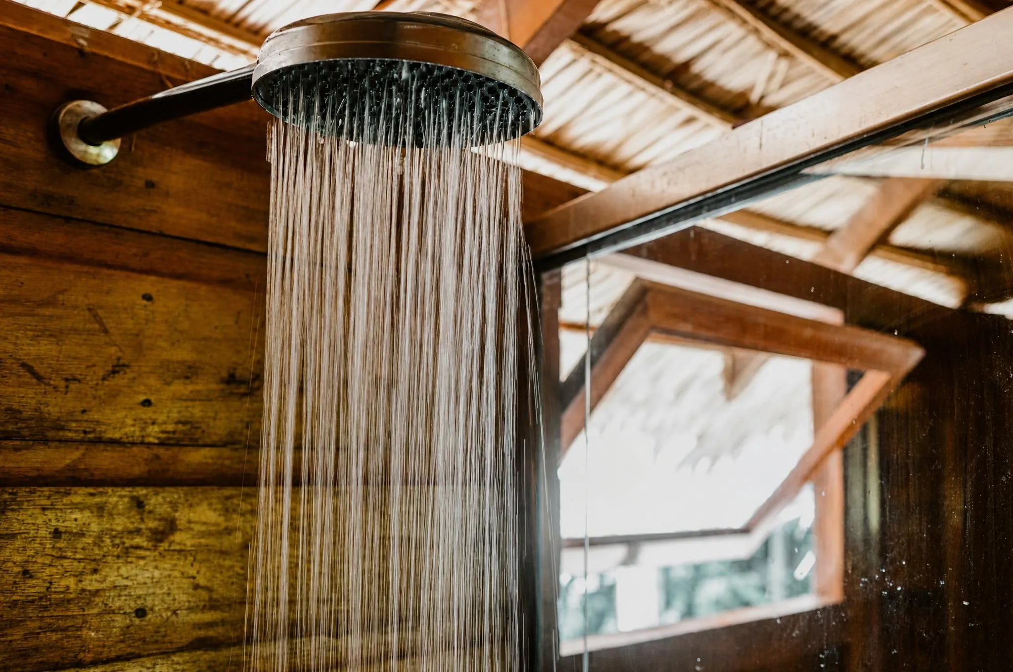 Chuveiro aberto num banheiro de madeira, a água caindo em fio contra a luz que entra pela janela