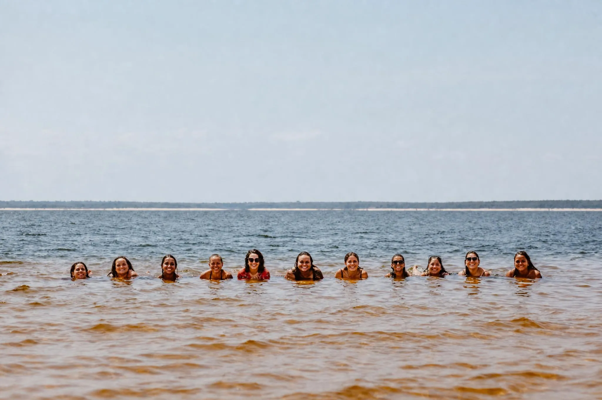 Fileira de onze mulheres lado a lado dentro da água rasa do rio, sorrindo de frente para a câmera