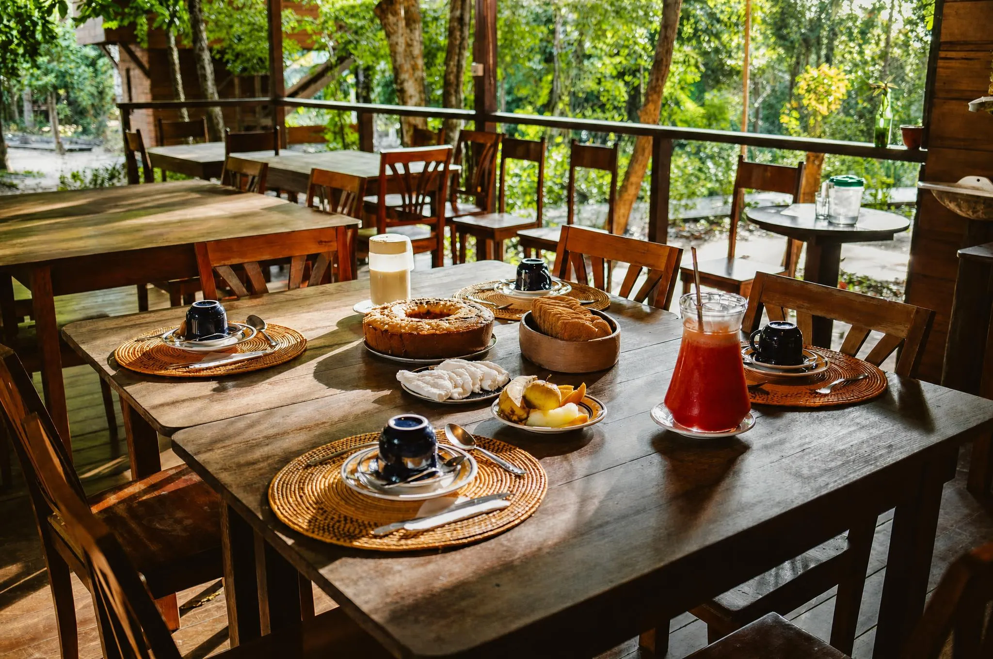 Café da manhã na mesa comprida de madeira: esteiras redondas trançadas, canecas escuras, bolo, pão, fruta fatiada e jarra de suco, aberto para a floresta