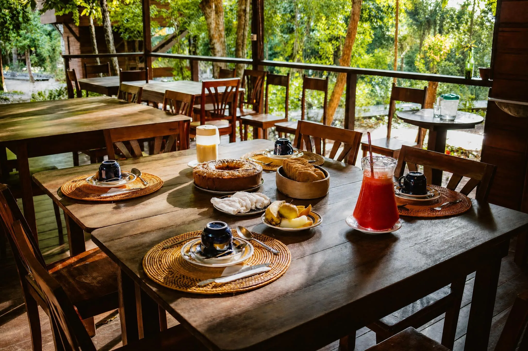 Mesa de café da manhã posta no pavilhão aberto: bolo, tapioca, fruta fatiada e jarra de suco vermelho, com a floresta atrás