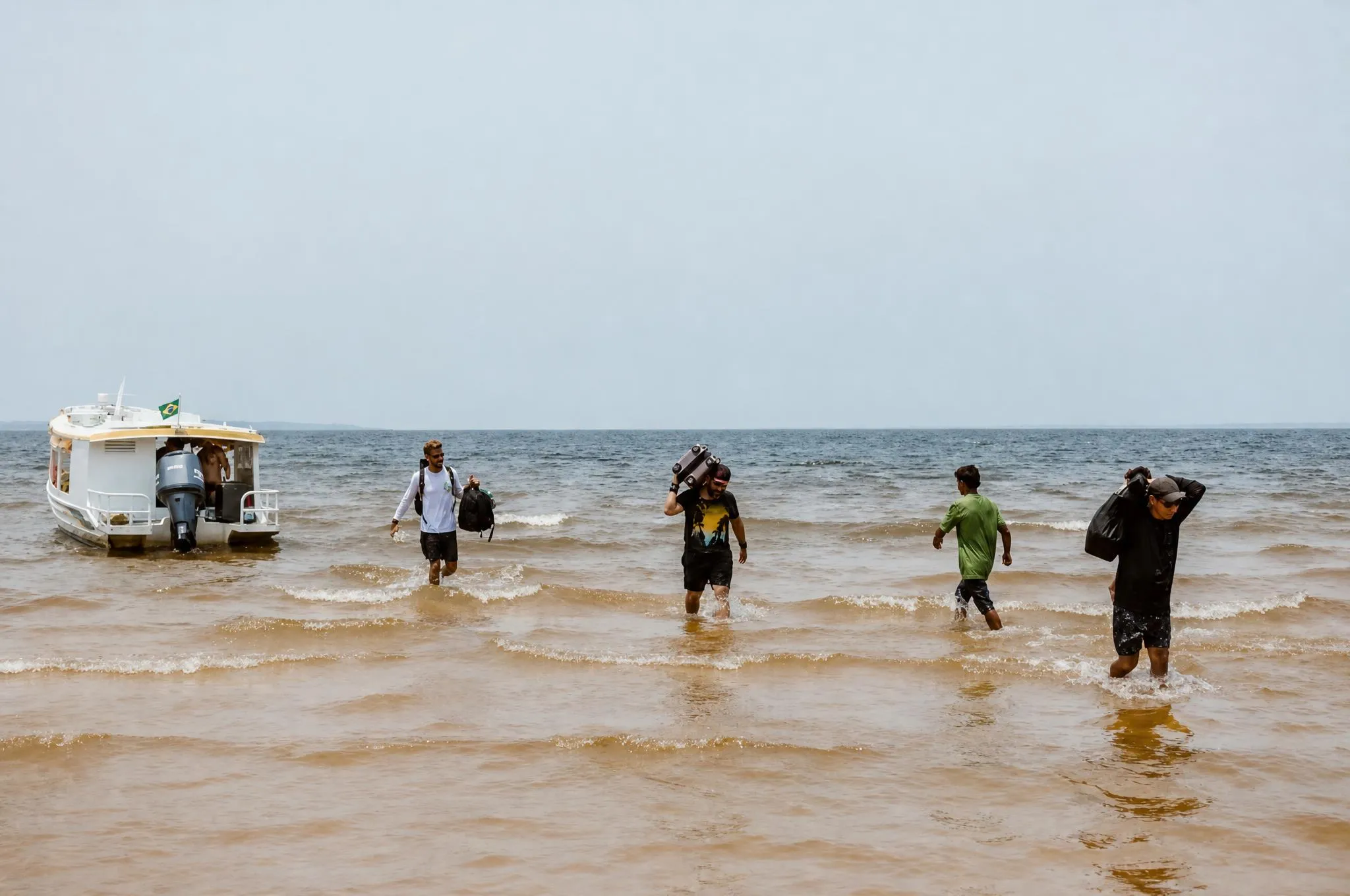 Pessoas desembarcando de um barco na água rasa junto à areia, com bagagem na mão