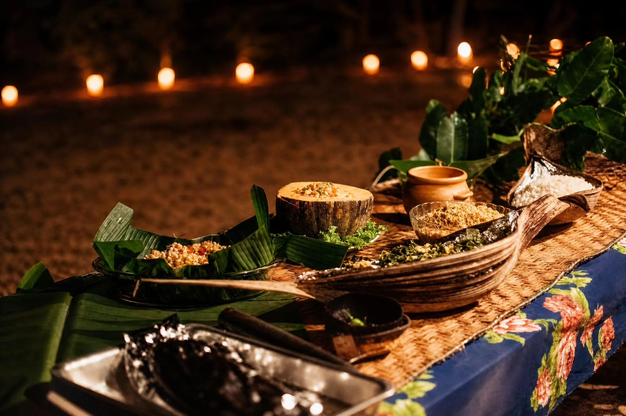 Mesa de jantar à luz de velas na praia: peixe, farinha e frutas servidos em folha de bananeira sobre esteira trançada