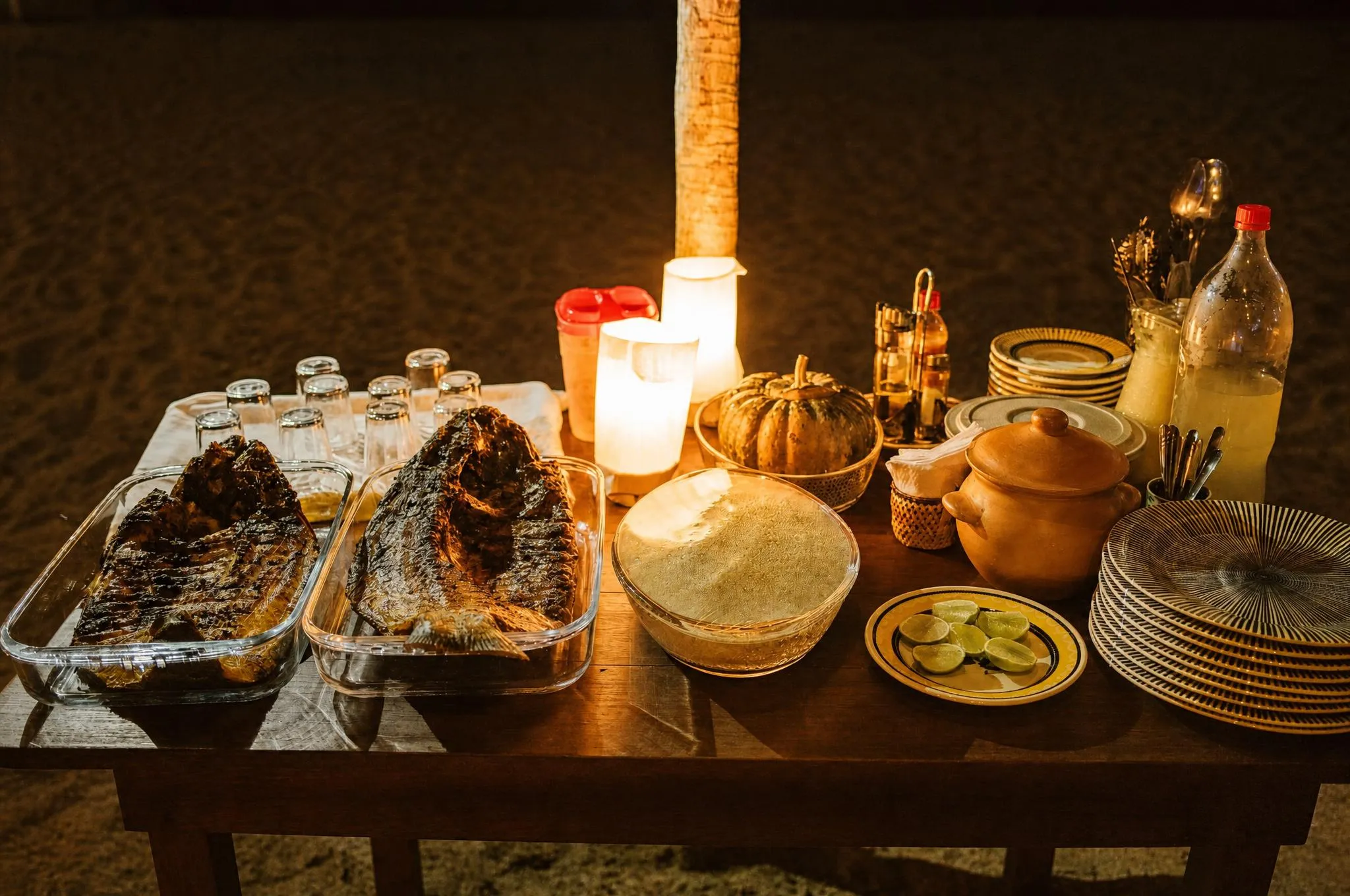 Peixes assados inteiros em travessas de barro à luz de velas, com farofa, limão e louça artesanal ao lado