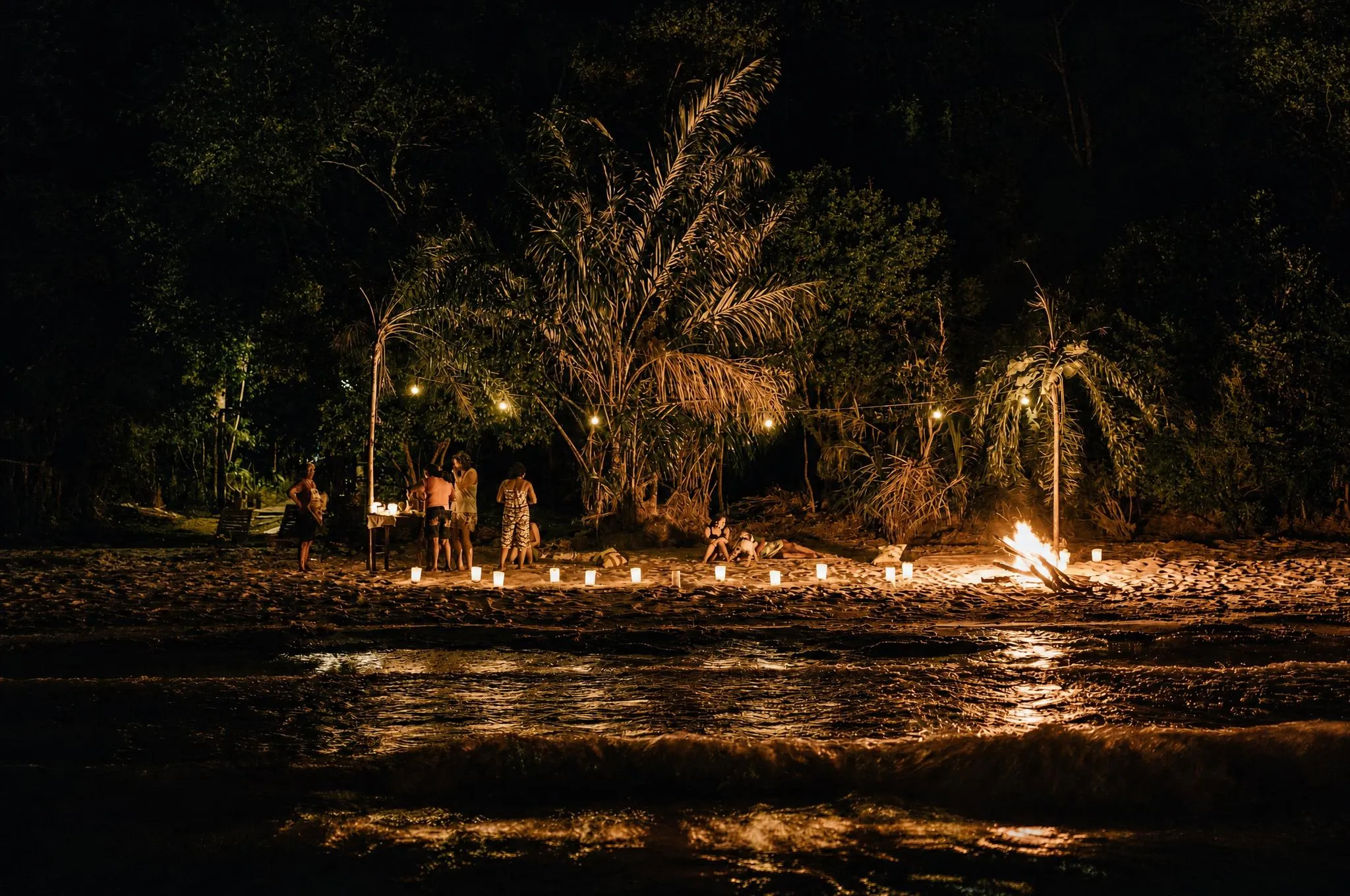 Praia à noite com fogueira acesa, fileira de velas na areia e hóspedes reunidos sob as palmeiras