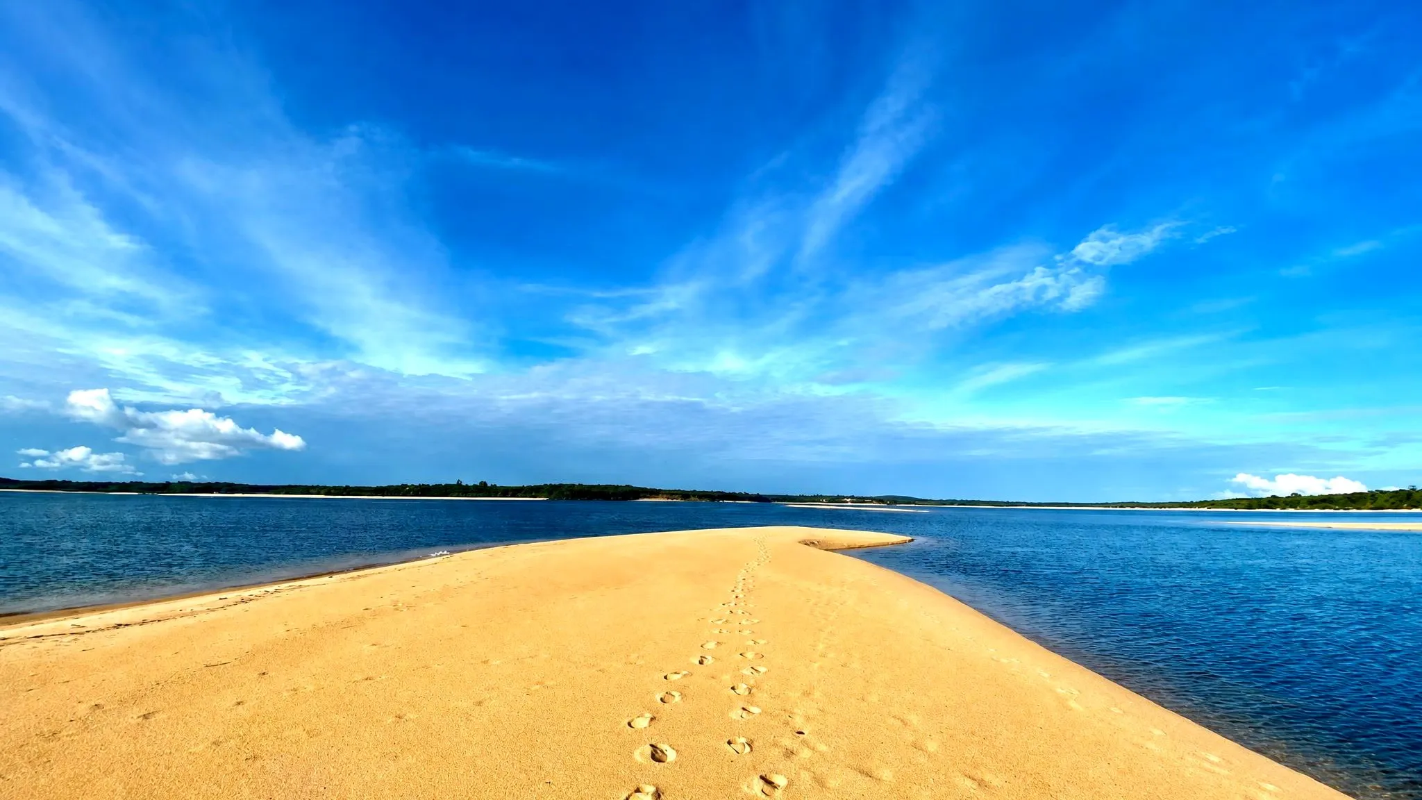 Banco de areia dourada avançando no rio sob um céu azul profundo com nuvens altas, com uma trilha de pegadas na areia