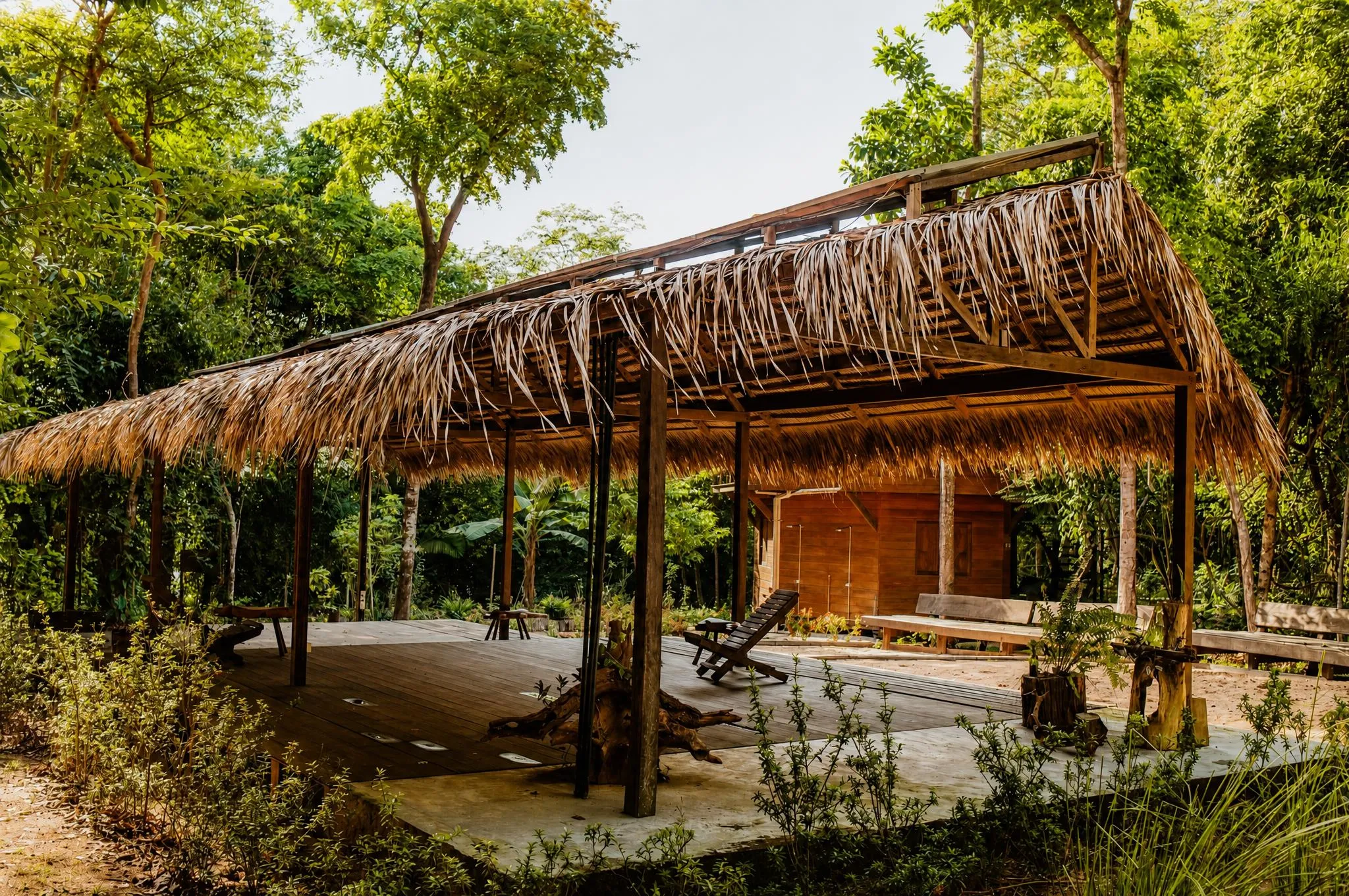 Pavilhão comprido de telhado de palha sobre deck de madeira, aberto para a mata, com bancos de tronco e espreguiçadeira embaixo