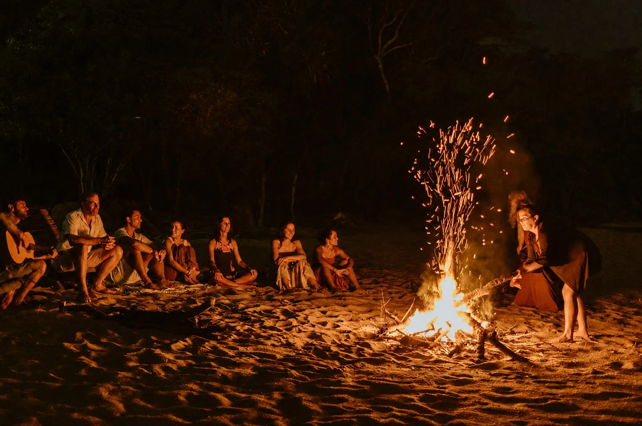 A circle of guests sitting on the sand at night around a fire, one of them with a guitar