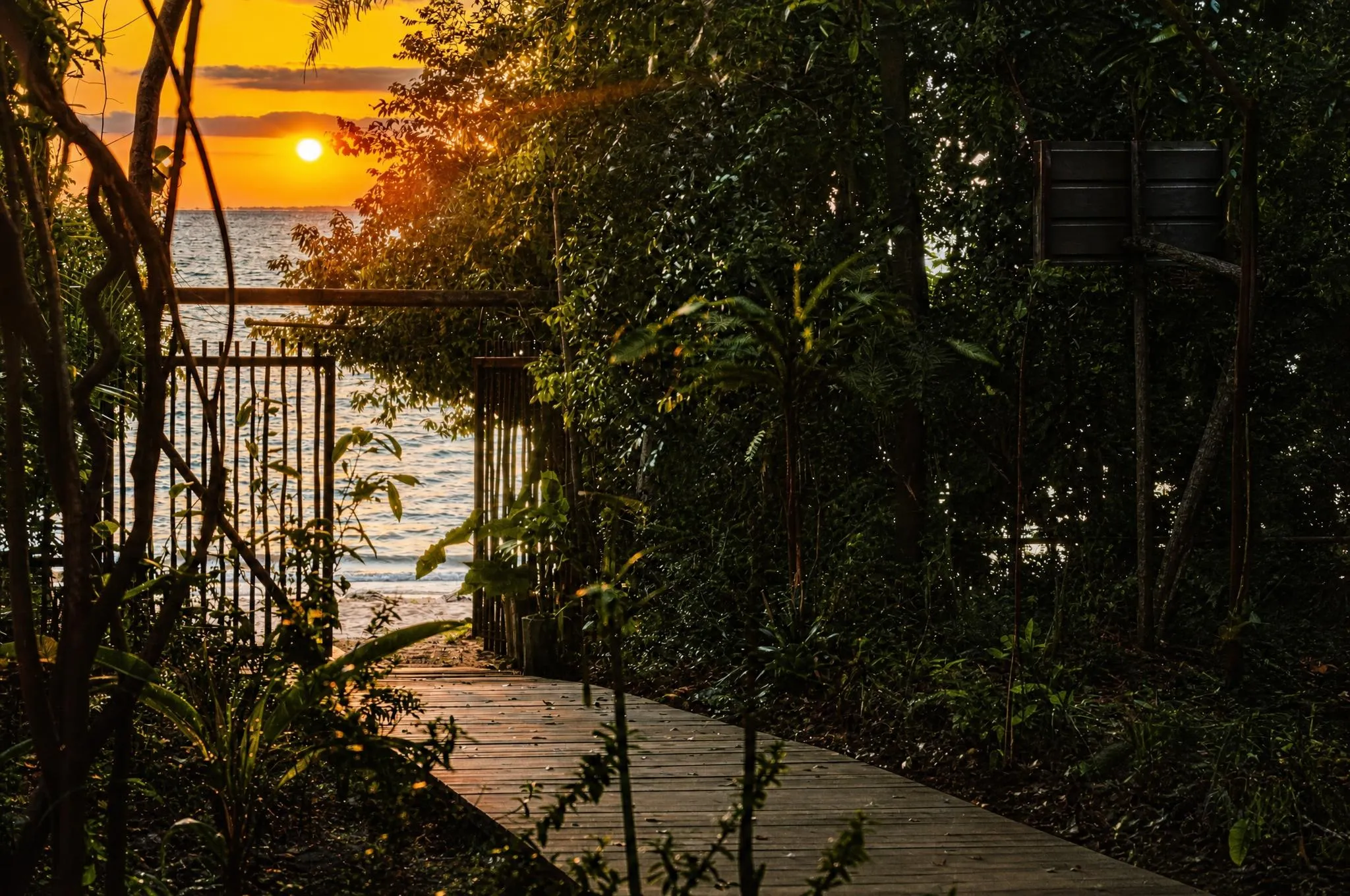 Sol baixo alaranjado sobre a água larga do rio, visto de uma passarela de madeira, com o portão e a vegetação em silhueta