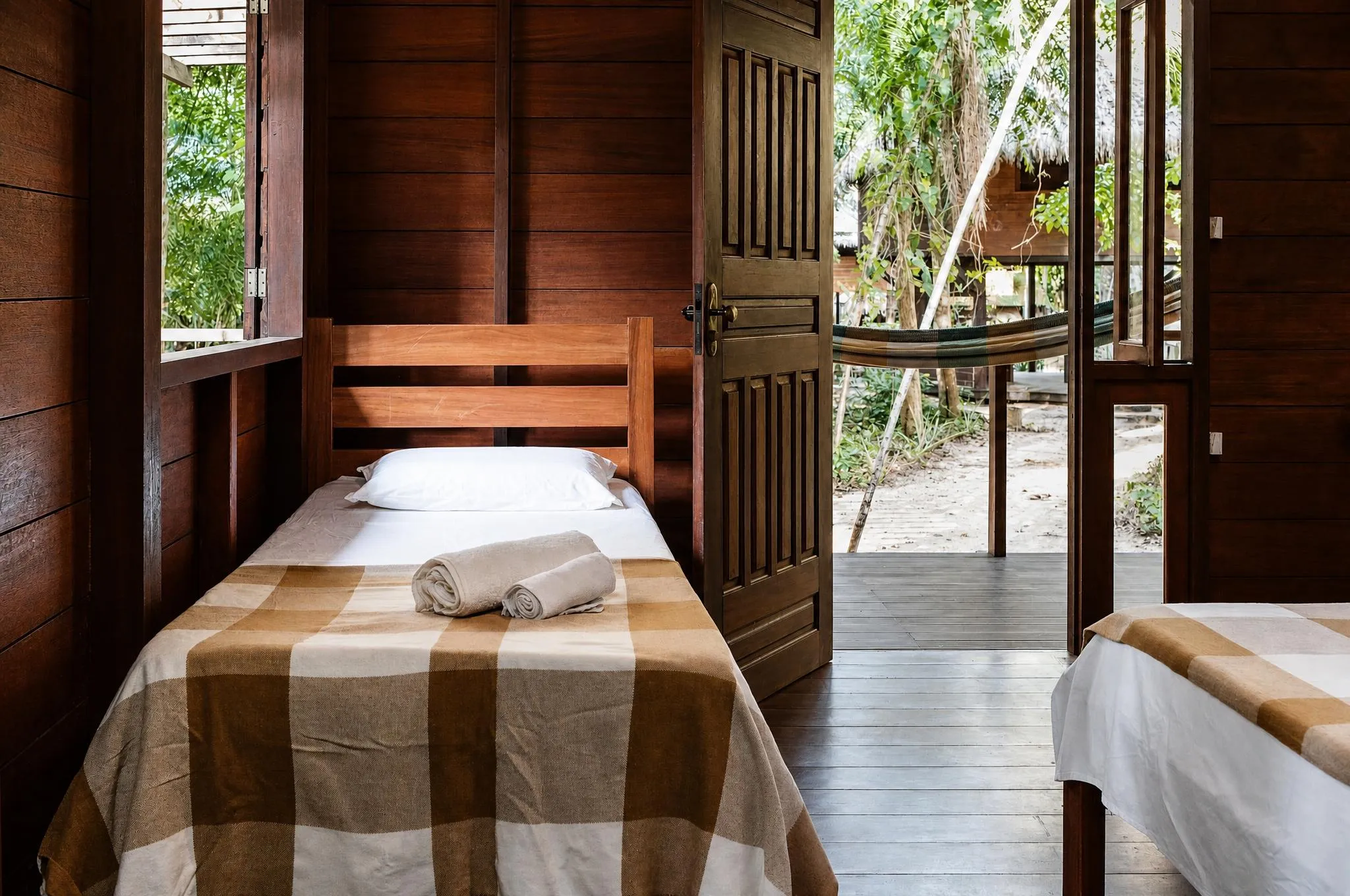 Quarto de paredes de mogno com cama de solteiro e manta xadrez, ao lado de uma porta de madeira de almofadas em relevo aberta para a varanda, onde há uma rede armada